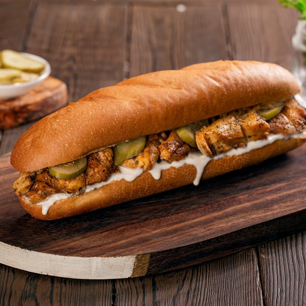 A chicken sub sandwich with pickles and white sauce on a wooden board, with a side of pickles.