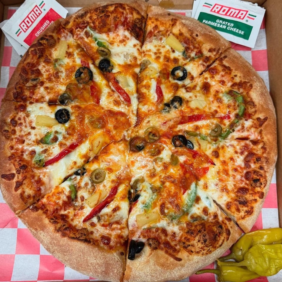 A Roma Pizza & Pasta pizza with olives, peppers, and pineapple, with parmesan and crushed pepper packets.