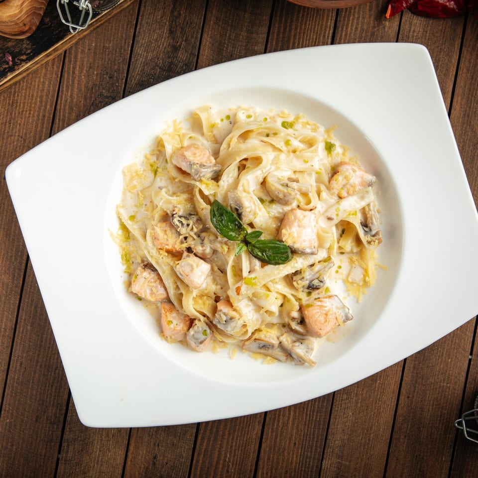 Creamy fettuccine pasta with salmon, mushrooms, and basil in a white bowl on a wooden table.