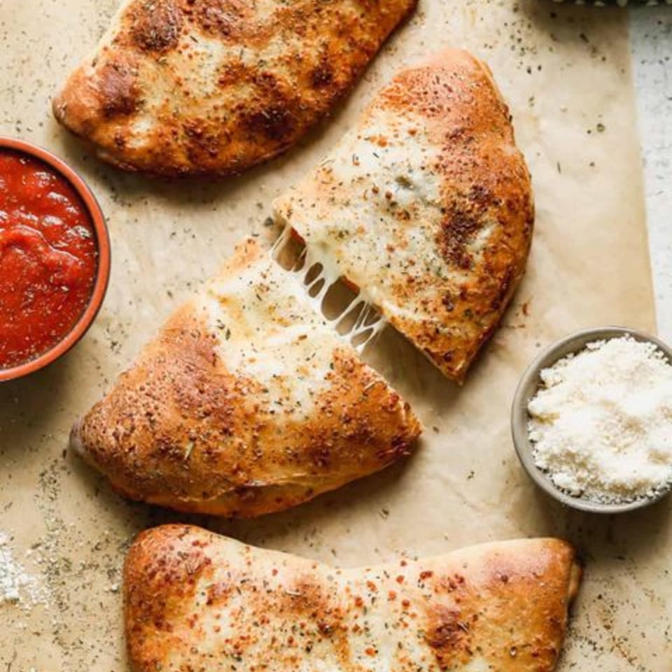 Three golden-brown calzones, one pulled apart with melted cheese strings, served with marinara and parmesan.